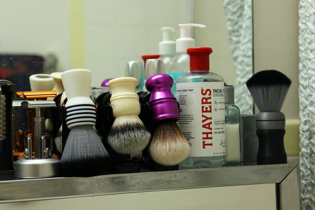 Multiple shaving brushes and skincare products on a bathroom shelf.